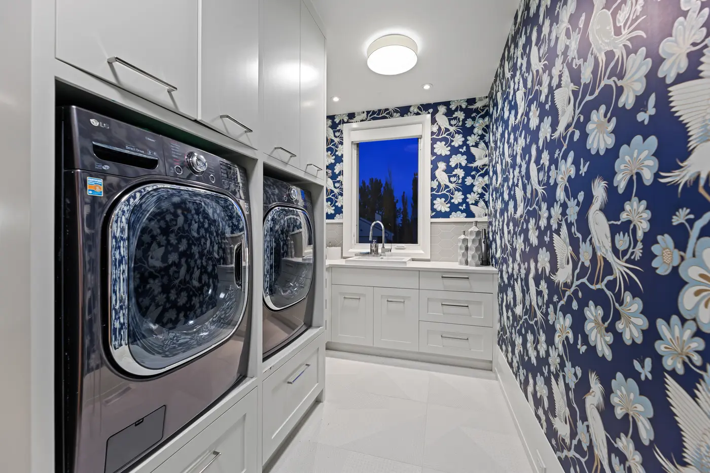A jewel-box laundry room wrapped in indigo heron-and-blossom wallpaper, with side-by-side graphite LG machines set into white cabinetry beside a hexagon-tiled sink wall and twilight window.