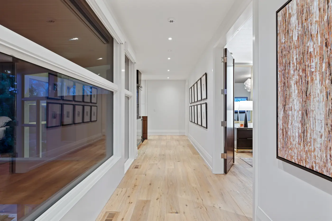 A light-filled upper corridor with white-oak floors, interior windows reflecting the photo gallery, framed print rows on the right and a bedroom with brass-trimmed nightstand glimpsed through a walnut door.