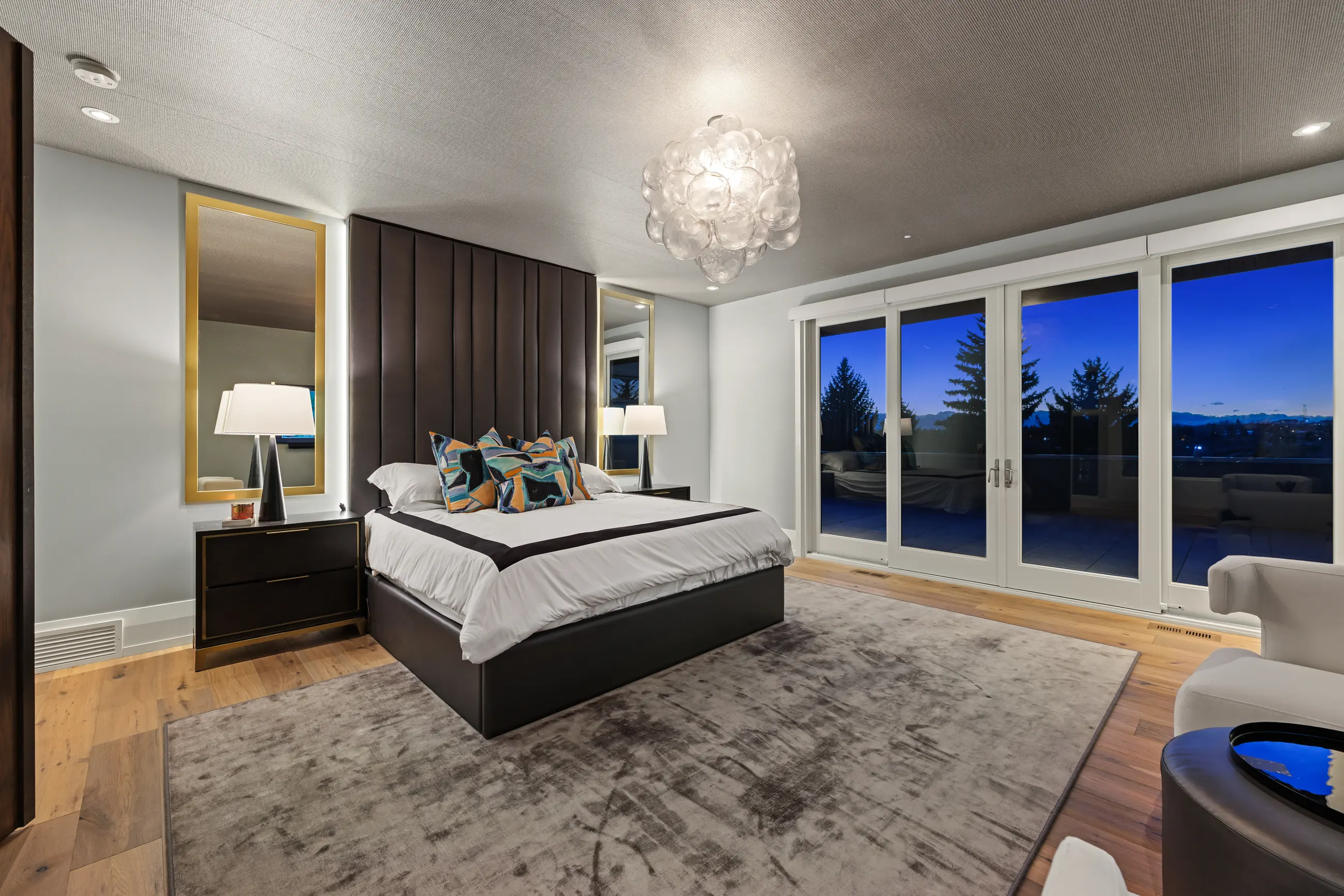 The primary bedroom at twilight, with a floor-to-ceiling channel-tufted leather headboard flanked by backlit brass mirrors, a bubble-glass chandelier and glass doors opening to a private terrace with treetop views.