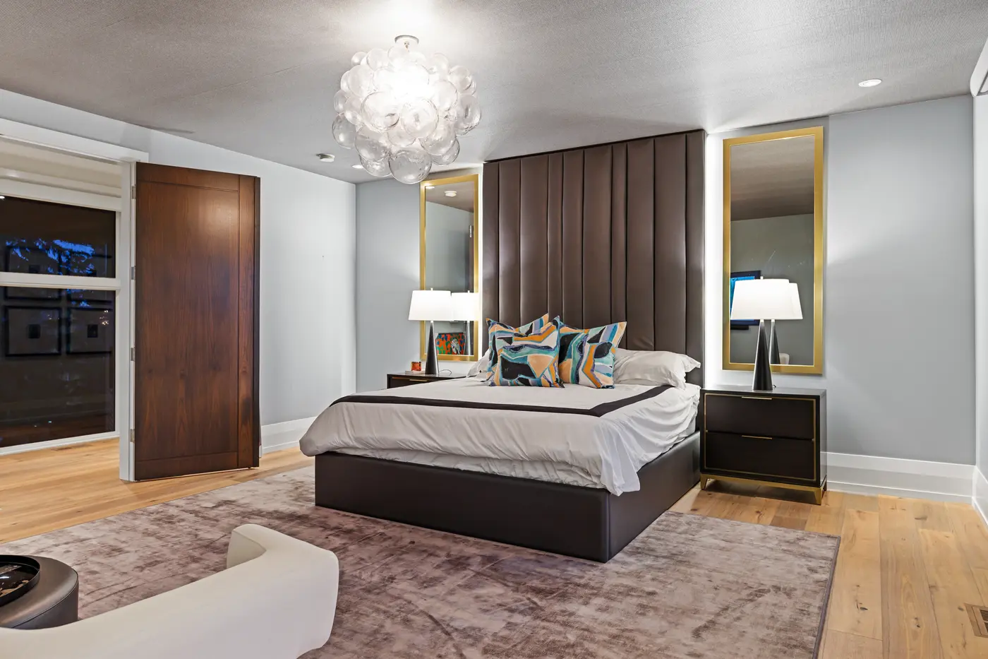 A wide view of the primary suite showing the chocolate leather feature headboard, brass-framed backlit mirrors over dark nightstands, a clear bubble chandelier and a walnut privacy door to the hall.