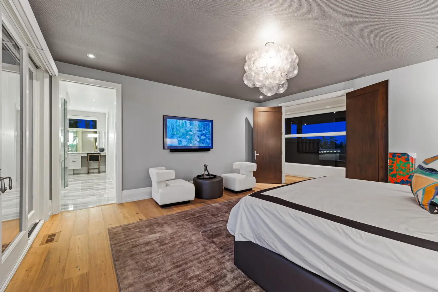 Looking across the primary bed toward a wall-mounted television with Monet water lilies, twin white lounge chairs, walnut double doors and the marble-floored ensuite beyond.