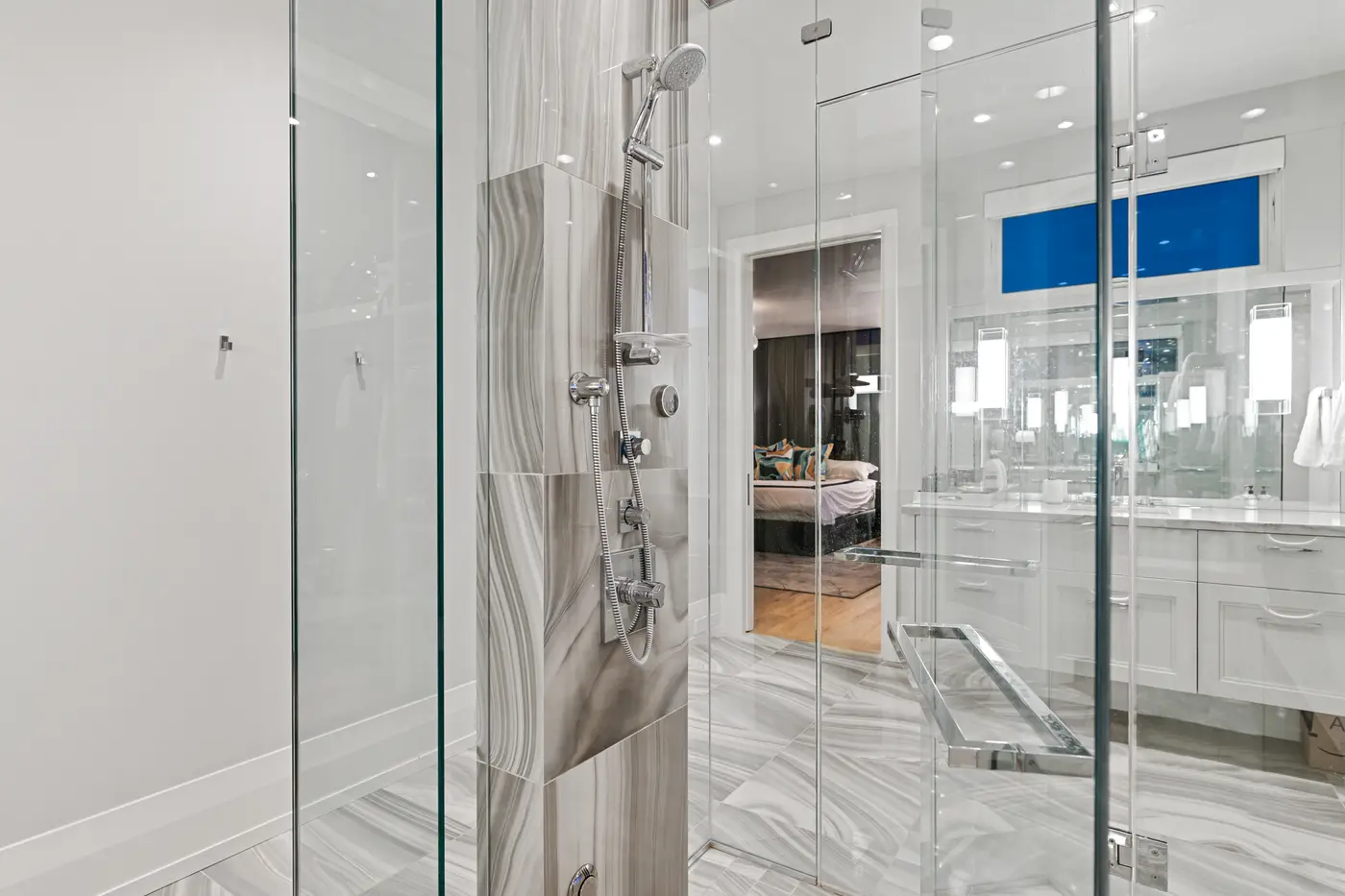 Inside the primary shower, polished chrome rain and handheld fixtures mount to a taupe agate-patterned tile column, glass panels reflecting the bedroom's leather headboard beyond.