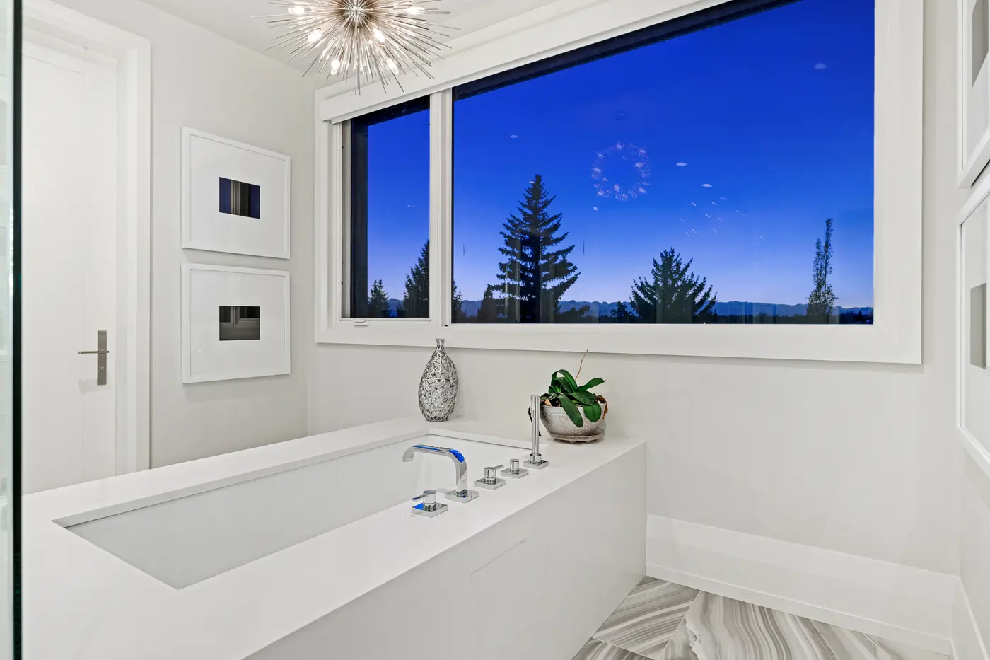 The primary soaker tub with deck-mounted chrome fittings set against a panoramic twilight window of silhouetted evergreens, a starburst chandelier sparkling overhead.