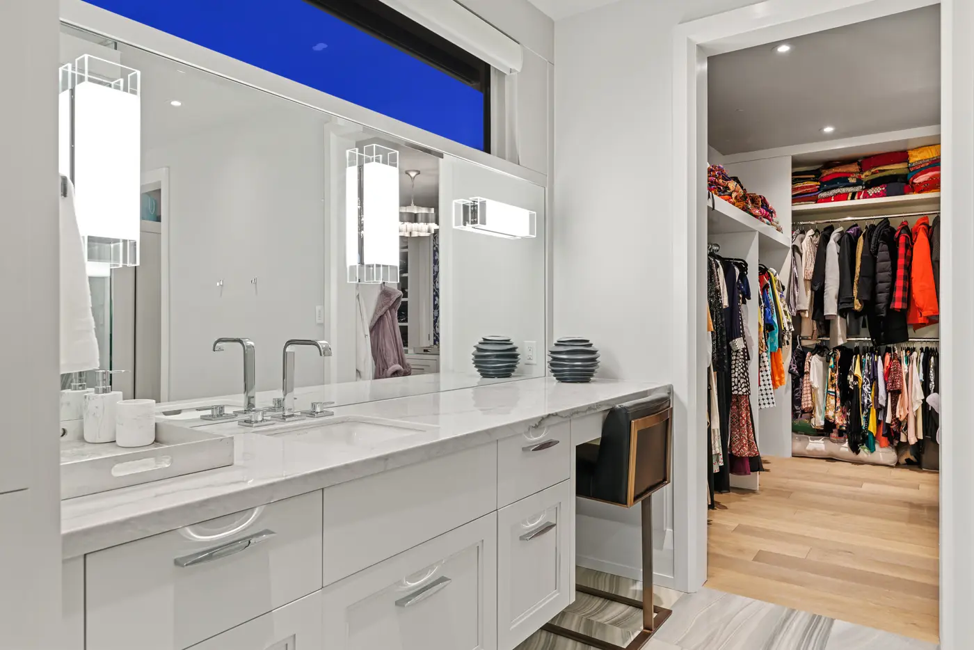 The marble-topped primary vanity with double chrome faucets and crystal sconces, its mirror band running beneath a clerestory window toward the color-filled walk-in wardrobe beyond.
