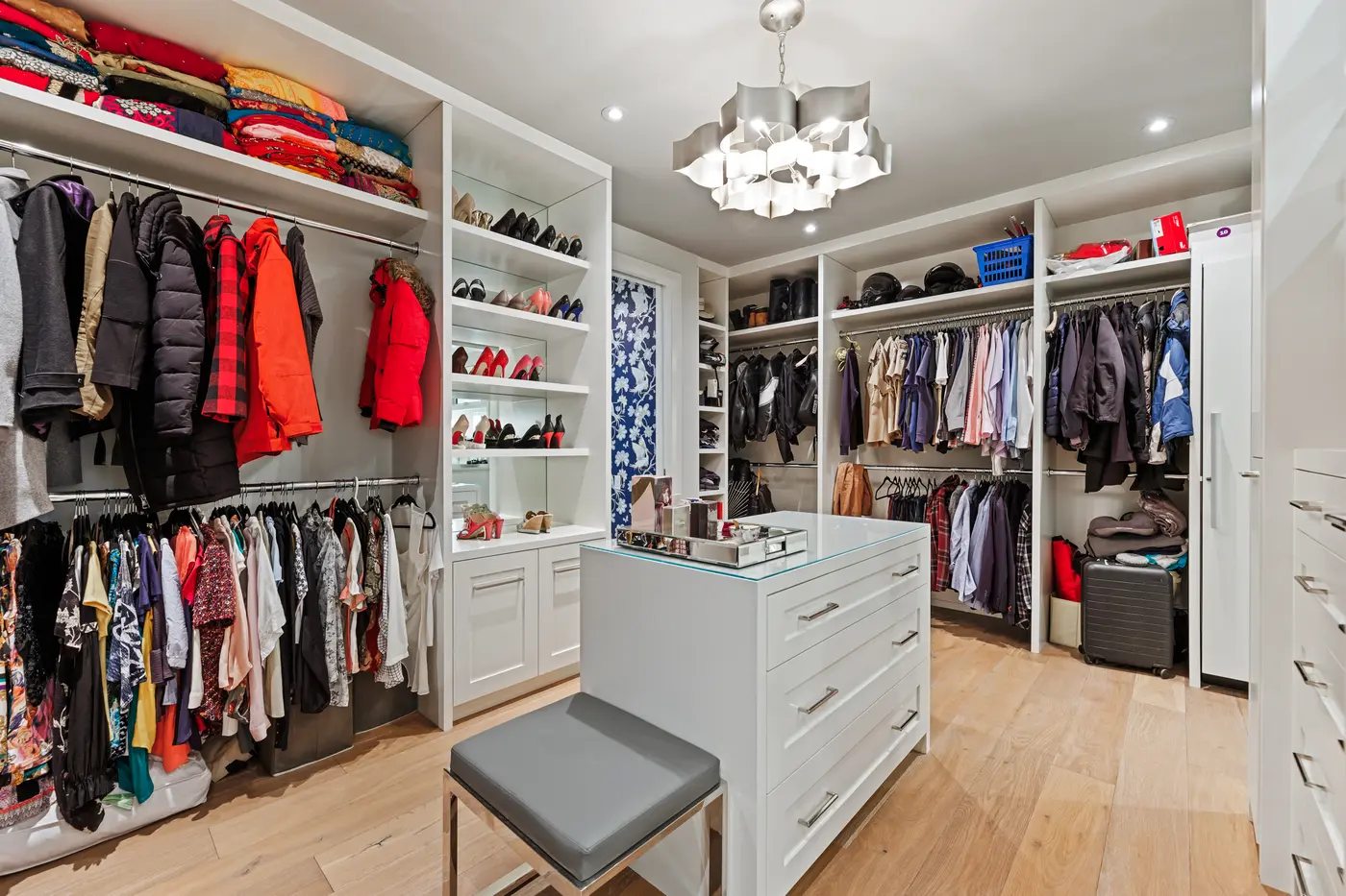 A boutique-style dressing room with a mirrored-top island, silver leaf chandelier, mirrored shoe shelving, and full-height wardrobe walls organized with coats, gowns and folded saris.