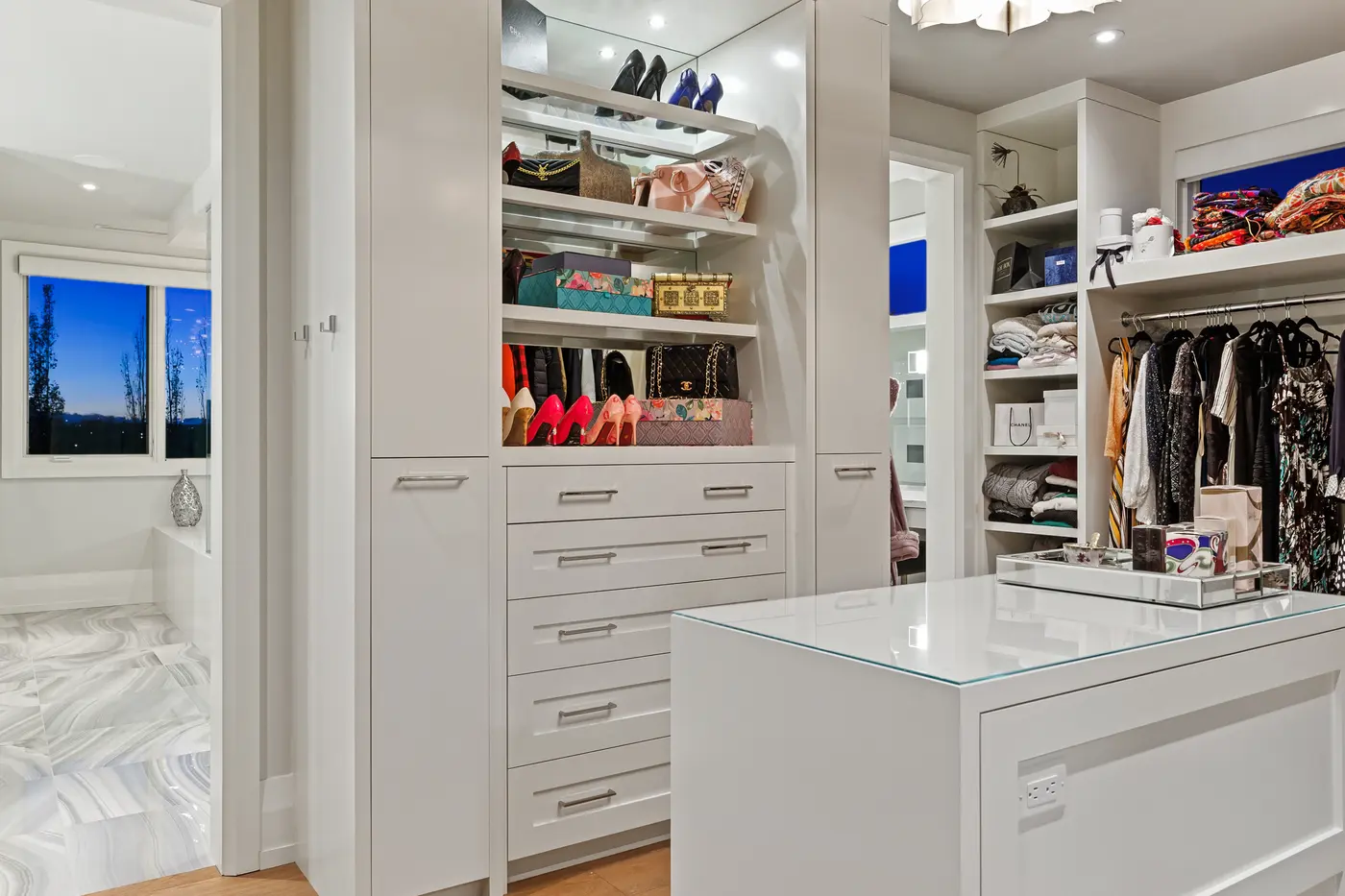 The dressing room's display wall of illuminated shelves styled with designer heels and handbags above a bank of white drawers, beside the glass-topped packing island and marble bath beyond.