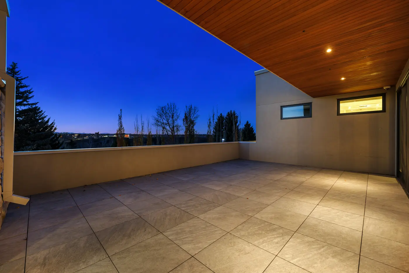 An expansive private roof terrace at deep dusk, its porcelain-tile deck wrapped by a solid parapet with glass wind screen and a warm cedar soffit overhead, city lights flickering on the horizon.