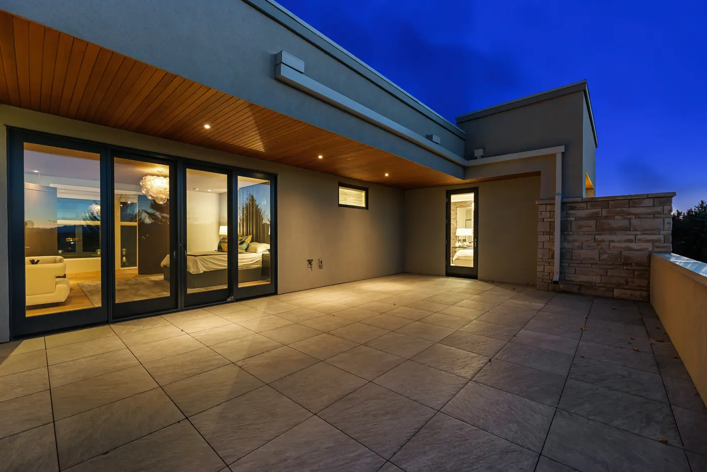 The primary suite's terrace at twilight, where glass doors reveal the lit bedroom and bubble chandelier, stone-capped parapet walls meeting the cedar overhang and porcelain pavers.