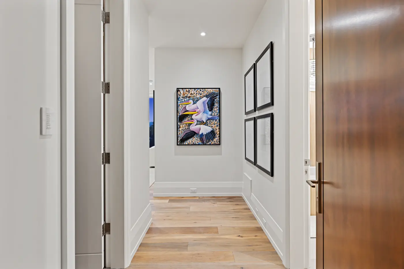 A crisp white hallway leading past a walnut door to a gallery moment: a vivid pelican canvas flanked by grids of black-framed photographs over pale oak floors.