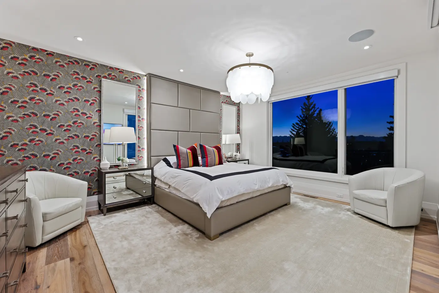 A guest bedroom with a feathered crimson-and-charcoal wallpaper feature wall, illuminated silver-framed mirrors, mirrored nightstands, and twin white barrel chairs facing a mountain-silhouette twilight window.