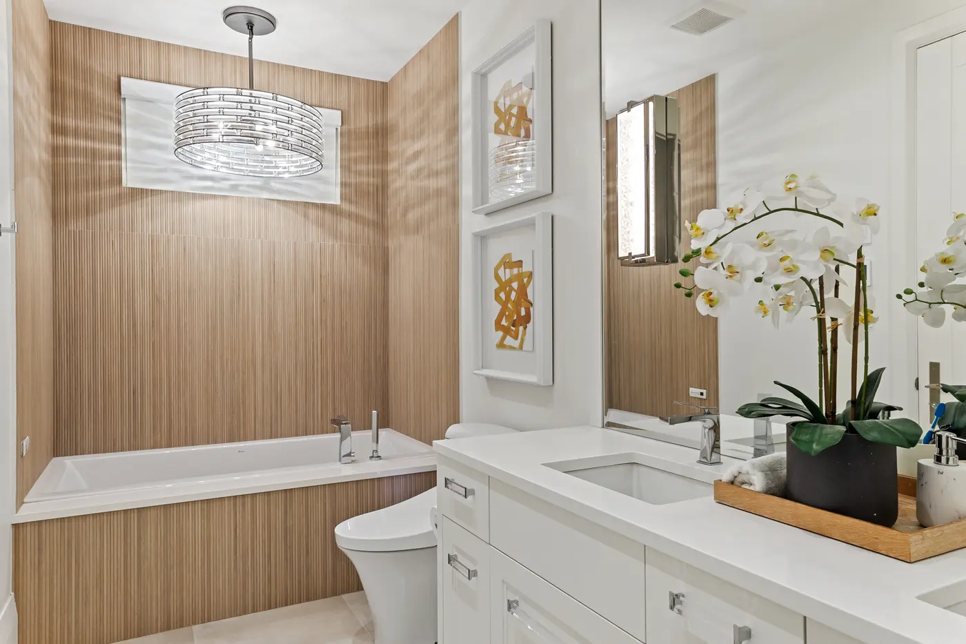A serene ensuite with a ribbed light-oak tile surround at the tub, a drum crystal pendant, white quartz vanity and an orchid styled on a wood tray beside the mirror wall.