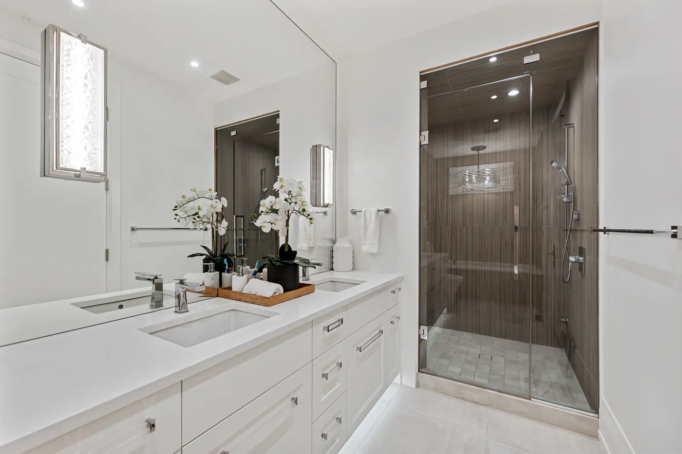 A white double-vanity bathroom with orchids and marble accessories reflected in a full-width mirror, facing a dark linear-tiled walk-in shower with handheld chrome fixtures.
