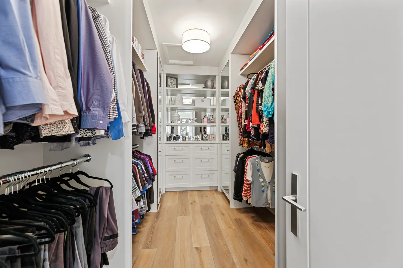 A walk-in closet with dual hanging levels of shirts and dresses, mirrored-back display shelving styled with perfume and decor, white built-in drawers and wide-plank oak floors.