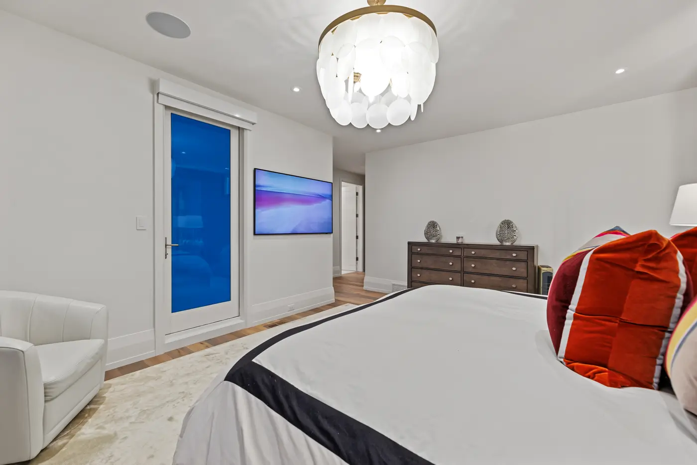 A guest bedroom viewed across the black-banded duvet toward a walnut dresser, wall-mounted television and paper-disc chandelier, with a twilight-blue glass door to the terrace.