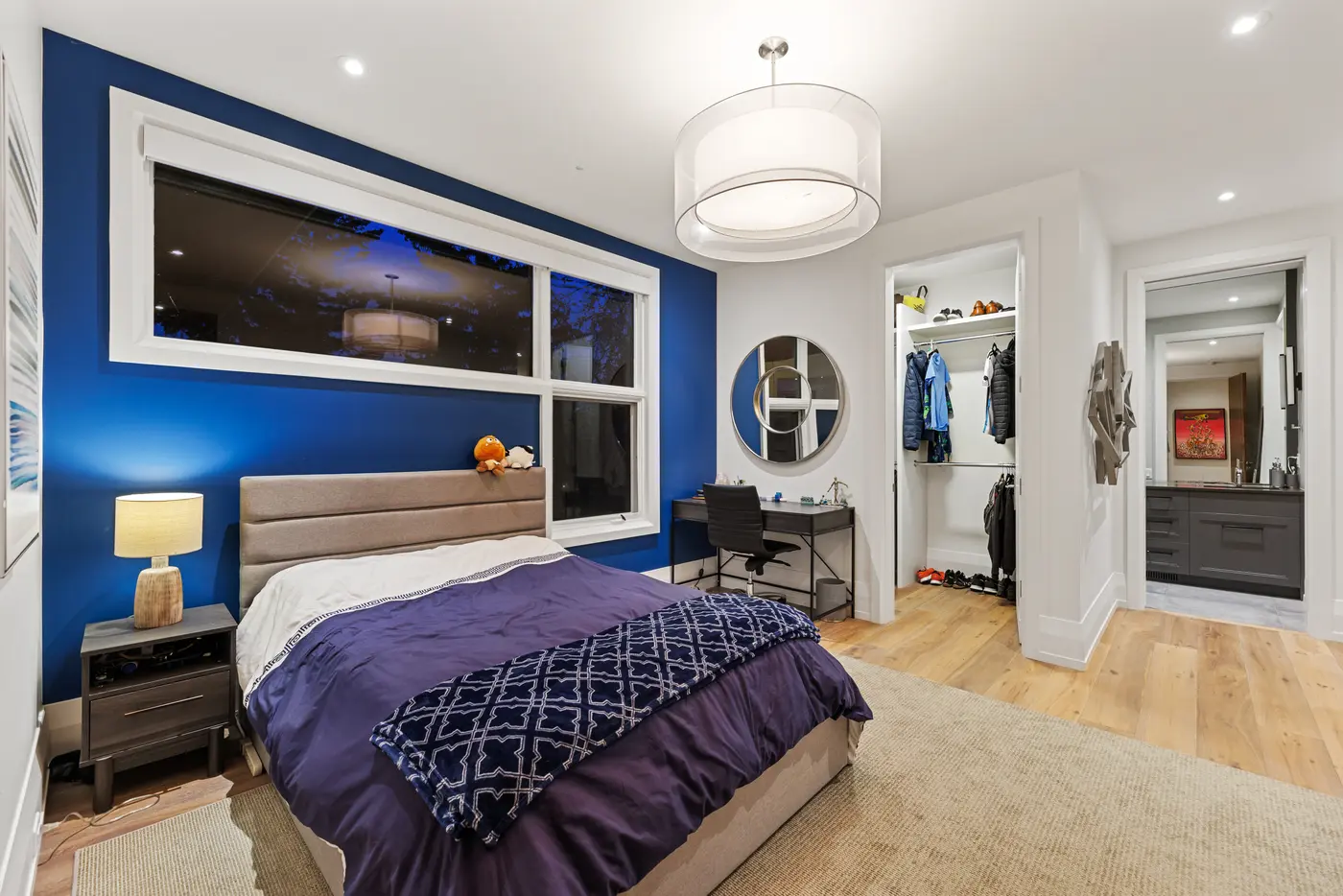 A boy's bedroom with a cobalt feature wall and corner windows, panel-tufted grey headboard, round mirror over a black desk, and a walk-in closet and dark-toned ensuite beyond.