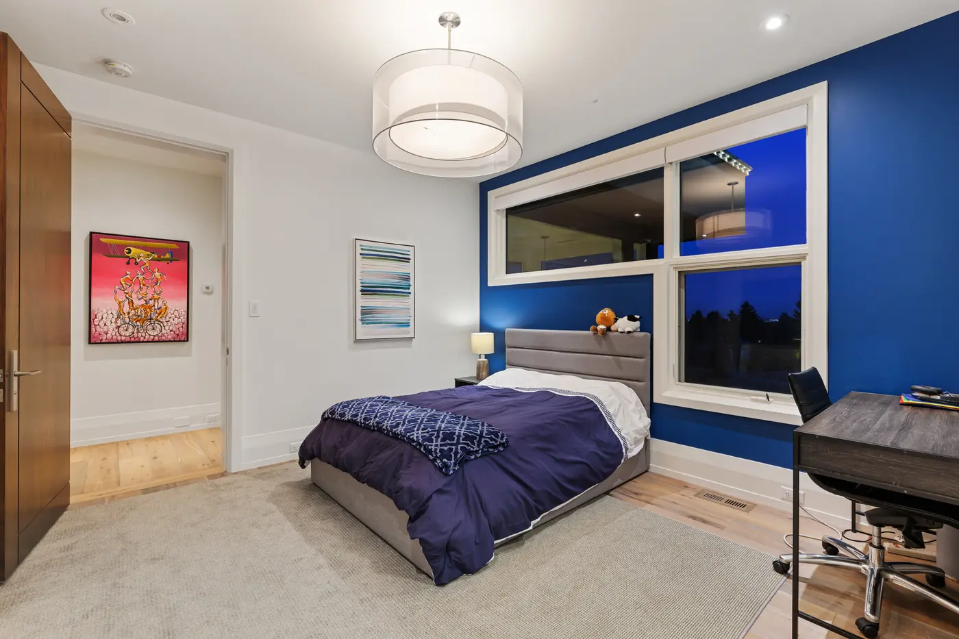Another angle of the cobalt bedroom showing the double-drum pendant, purple quilted bed with plush toys on the headboard, art-lined hall entry and dark wood desk at the window.