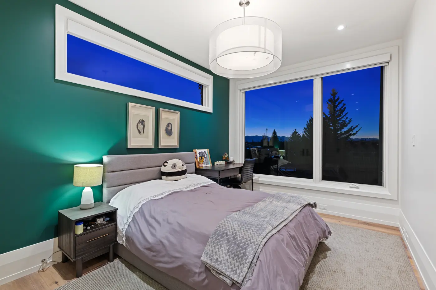 An emerald-walled bedroom with a grey channel headboard, panda plush on lavender bedding, agate prints and a broad picture window framing evergreens against the darkening sky.