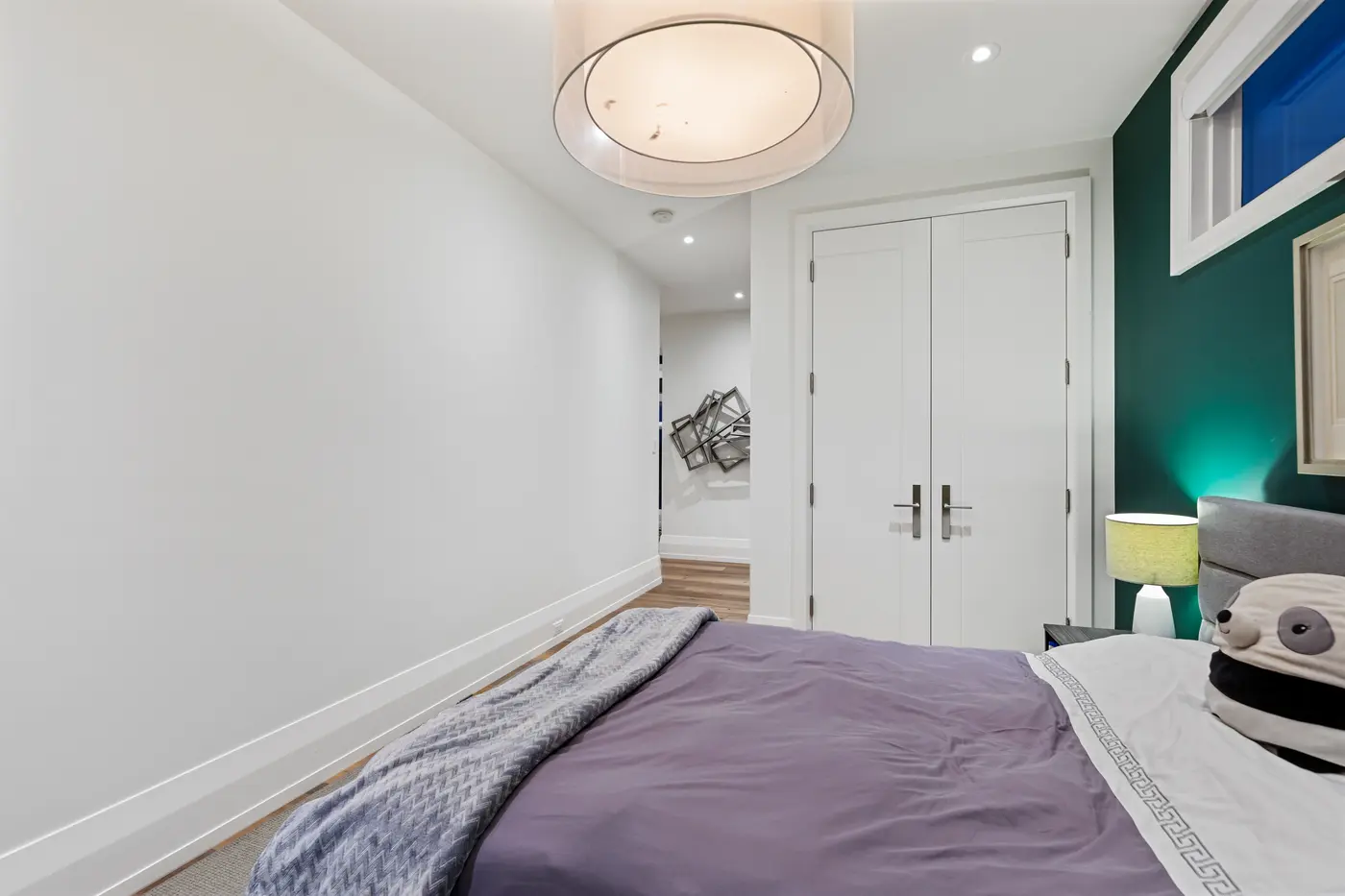 The emerald bedroom's entry view past a sheer drum pendant to white double closet doors and a hallway with sculptural metal wall art, lavender bedding in the foreground.