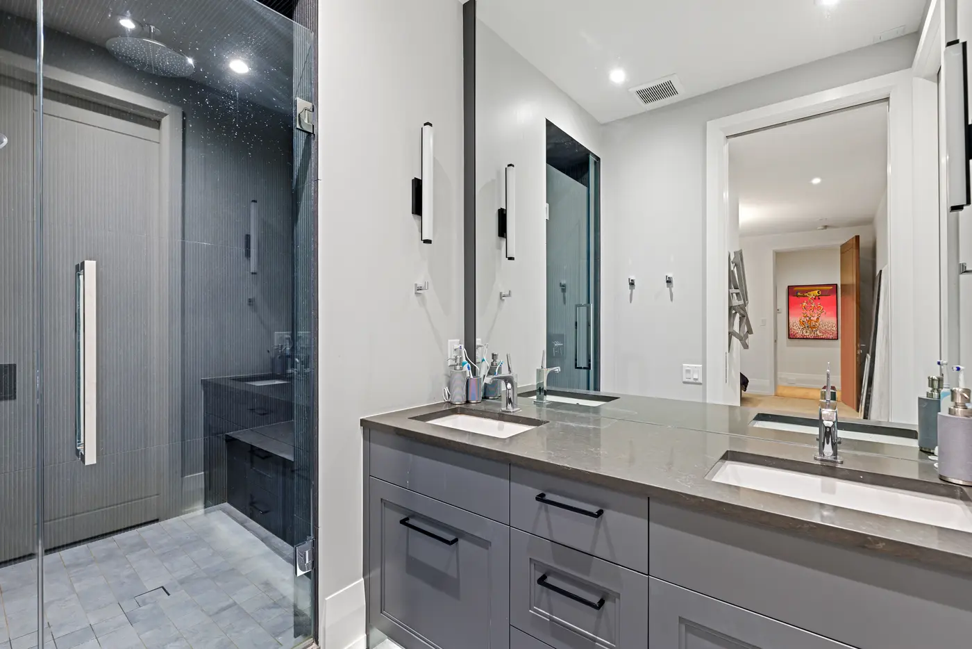 A grey-toned ensuite with double undermount sinks in a smoky quartz counter, matte-black hardware, backlit sconces and a rain-head shower wrapped in charcoal ribbed tile.