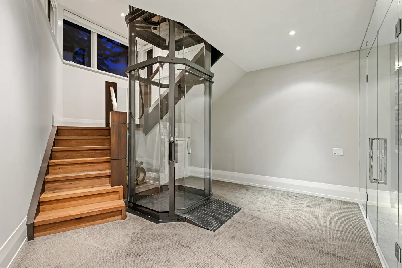 The glass elevator arrives at the carpeted lower level beside walnut-railed oak stairs and a full-height glass wall, twilight windows glowing above the landing.