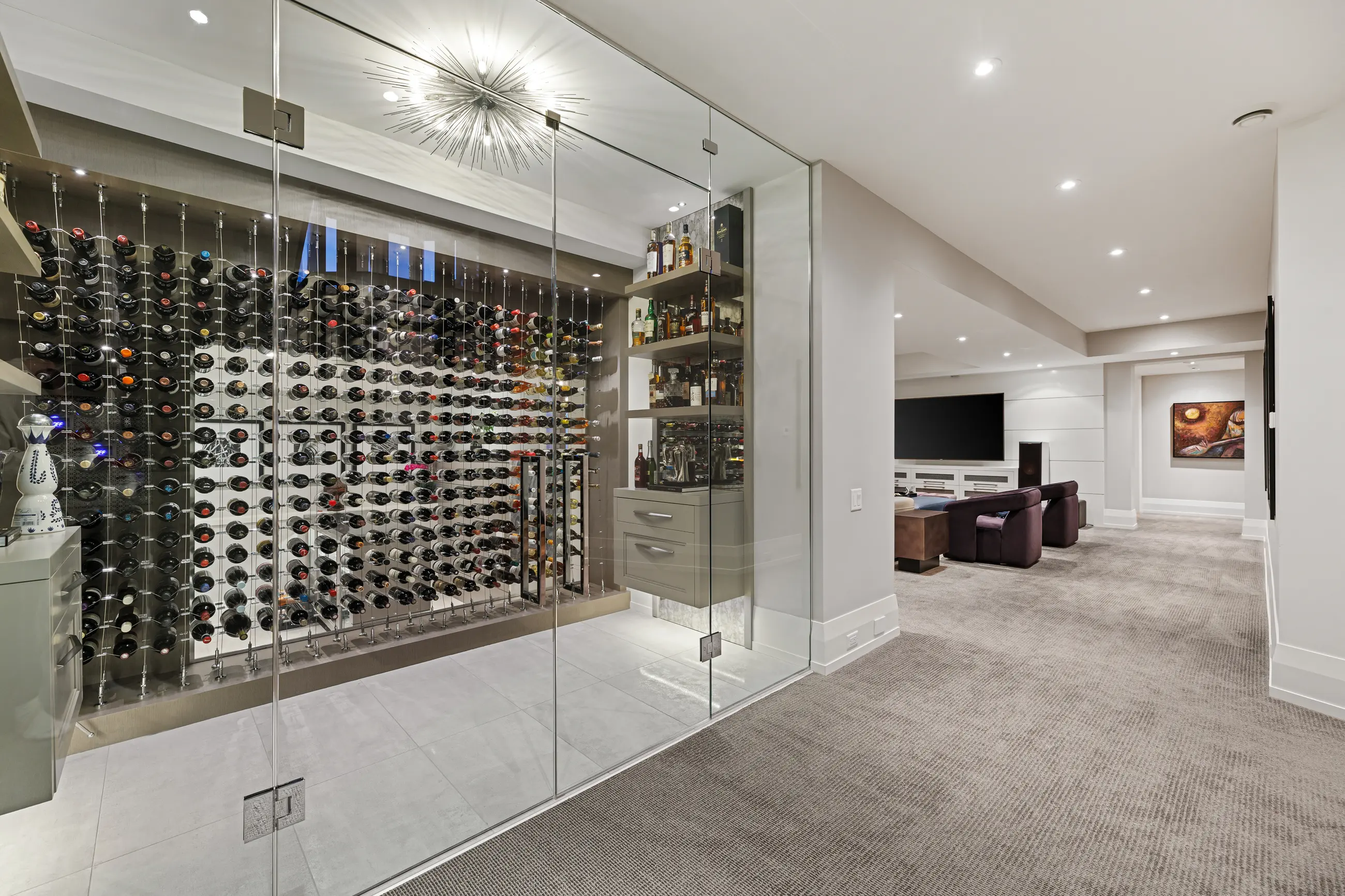 A glass-enclosed climate wine room displaying hundreds of bottles on cable-tension racks beside backlit whisky shelving, with the media lounge and its purple velvet chairs beyond.