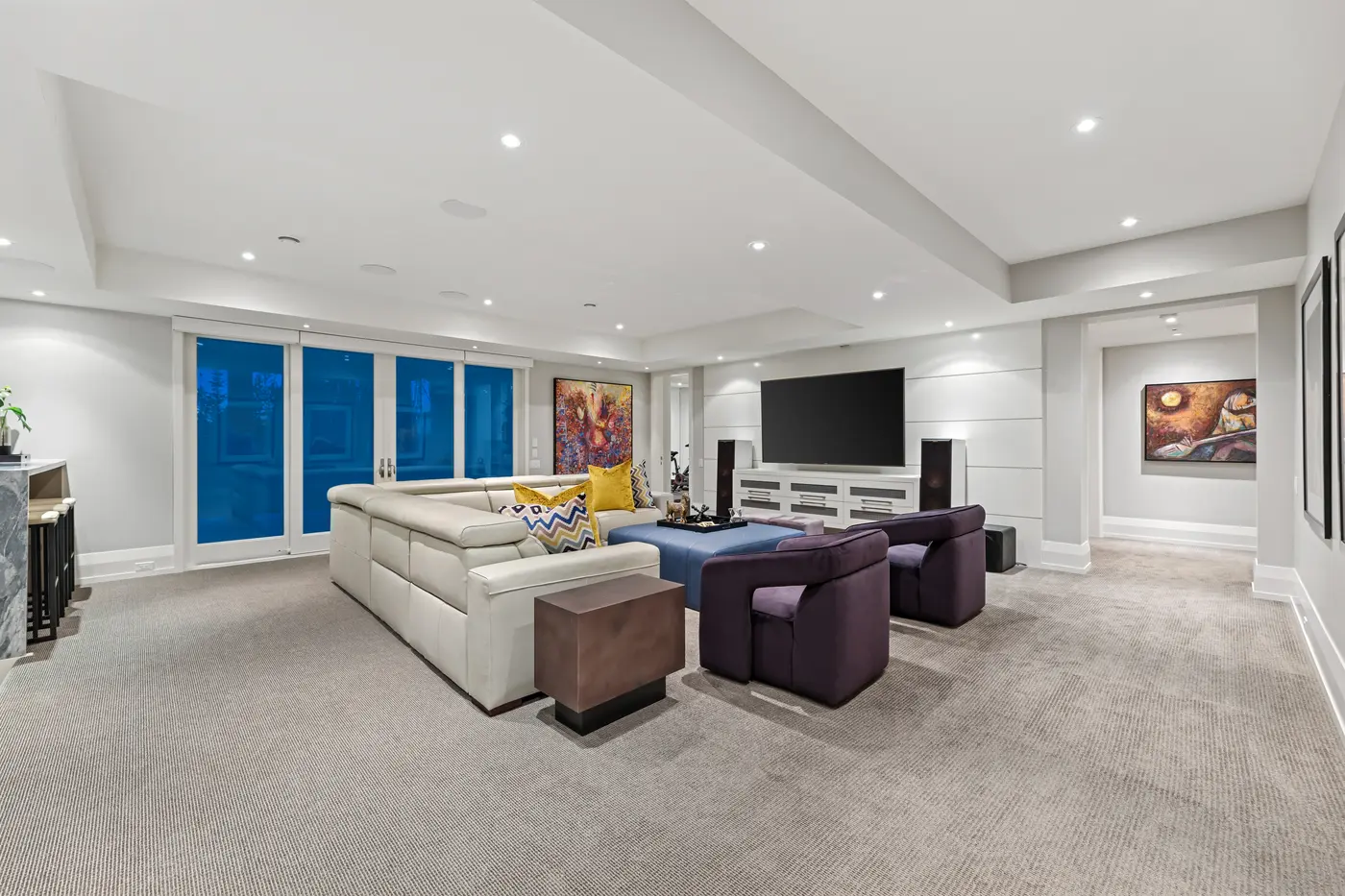 The lower-level media lounge with a cream leather sectional, plum velvet swivel chairs around a blue leather ottoman, wall-mounted screen with tower speakers and vibrant abstract canvases.