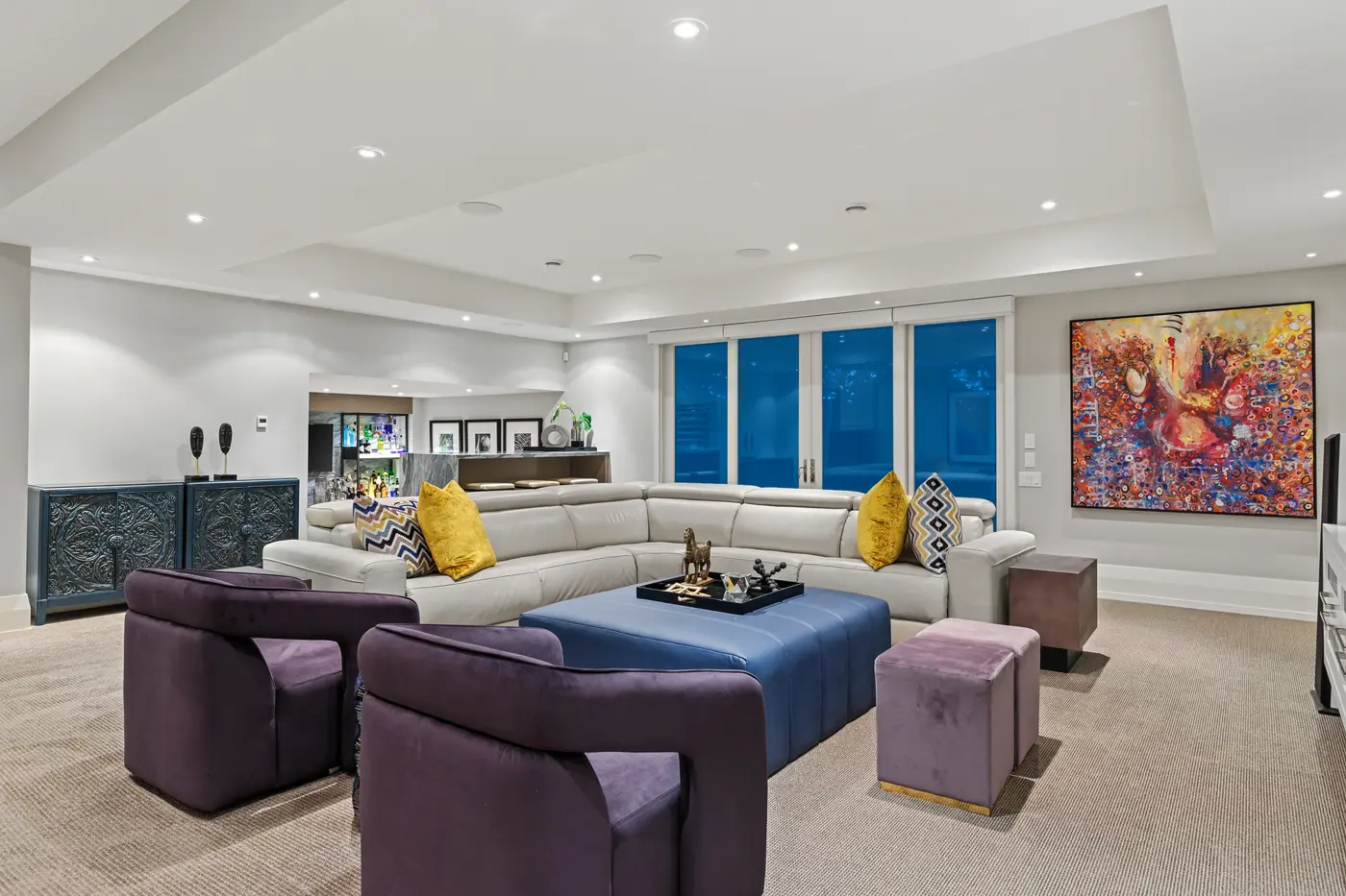 A wide view of the entertainment lounge showing the sectional and ottoman grouping, glass doors glowing blue at dusk, carved teal cabinets and a fiery abstract painting on the far wall.