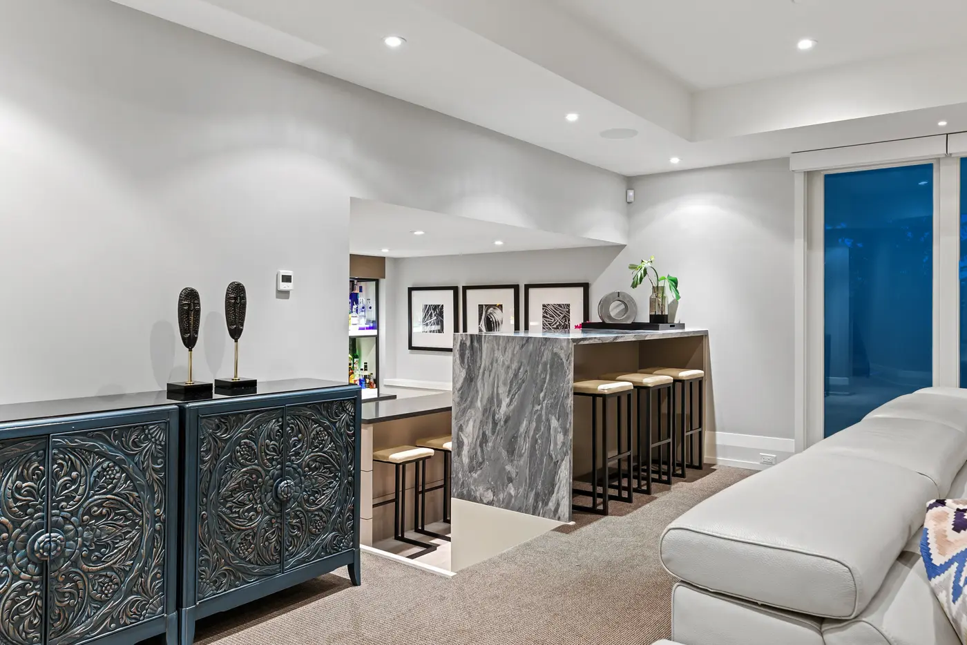 The lower-level bar's grey marble waterfall counter with leather-topped stools, flanked by intricately carved teal cabinets with sculptural masks and backlit spirits shelving beyond.