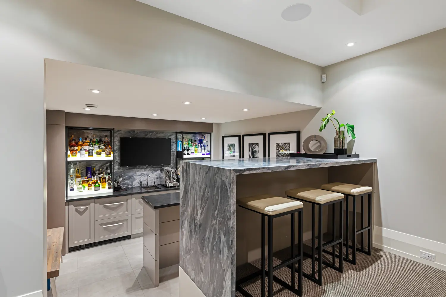 The home bar in full: illuminated glass-front spirit displays on a storm-grey marble backsplash, espresso machine and sink, television, and a dramatic marble waterfall island with three stools.