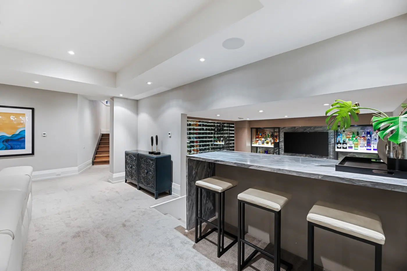 Looking across the bar's marble counter toward the glass wine wall and glowing liquor displays, cream barstools lined along the front and a monstera adding a fresh green accent.