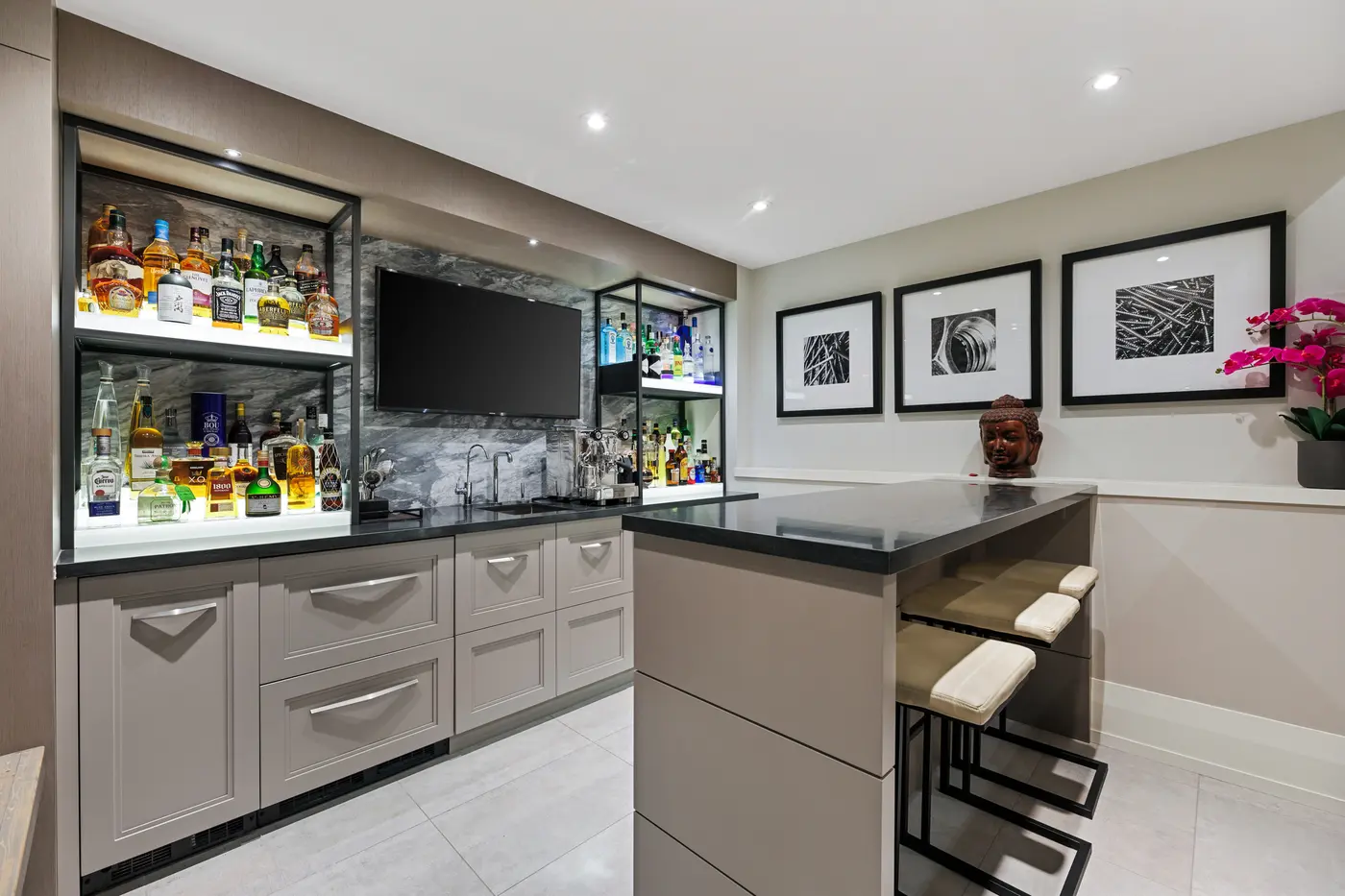 A close view of the bar's back counter with LED-lit shelves of whisky and tequila, black granite work top with prep sink and espresso machine, and black-and-white photography beside a Buddha bust.