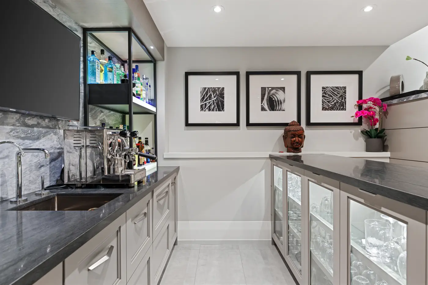 The lower-level bar's coffee and cocktail station, where a chrome espresso machine and backlit spirits shelving sit against dramatic grey marble beneath glass-front cabinets of glassware.