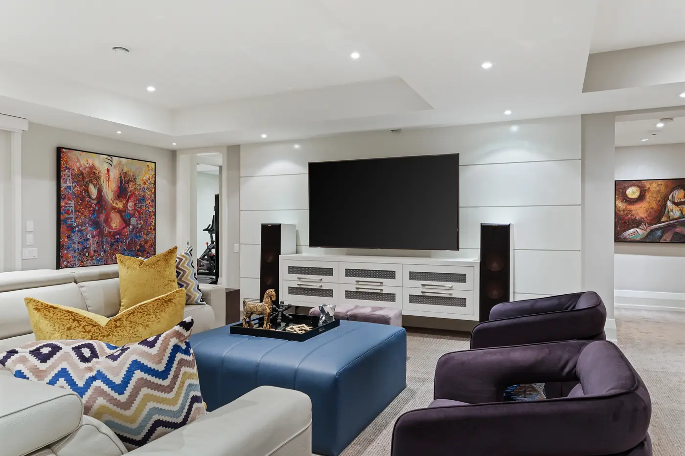 A wide media wall with floating console and reference tower speakers faces the lower level's leather sectional, layered with jewel-toned velvet chairs and a bold blue ottoman.