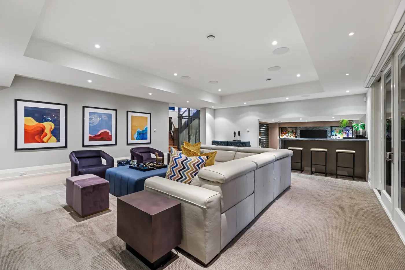 The full sweep of the walkout lower level, from the leather sectional and colourful triptych to the marble-topped bar with stools and the glass elevator beyond.