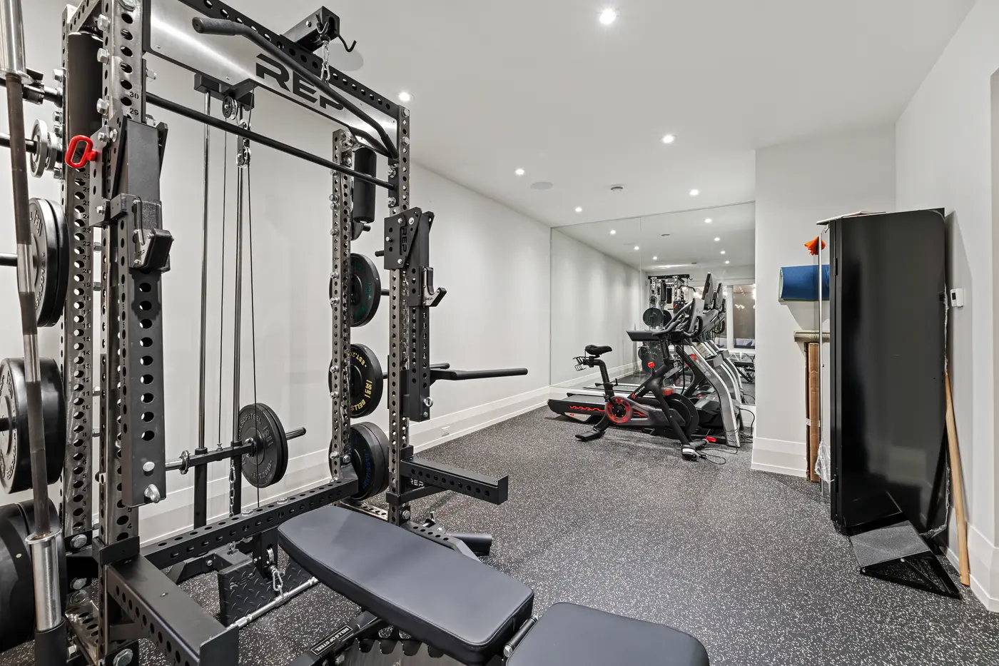The rubber-floored home gym is outfitted with a REP power rack, bumper plates, Peloton bike and treadmill against a full mirrored wall.