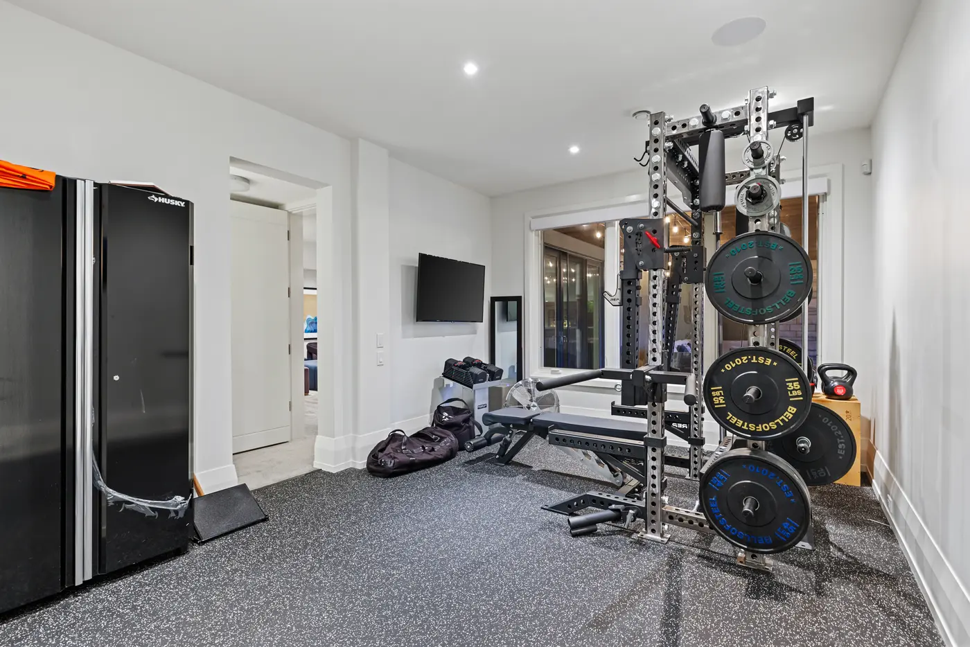 A second angle of the gym showing the loaded half rack, adjustable bench, kettlebells and wall-mounted television beside an interior window to the lower level.