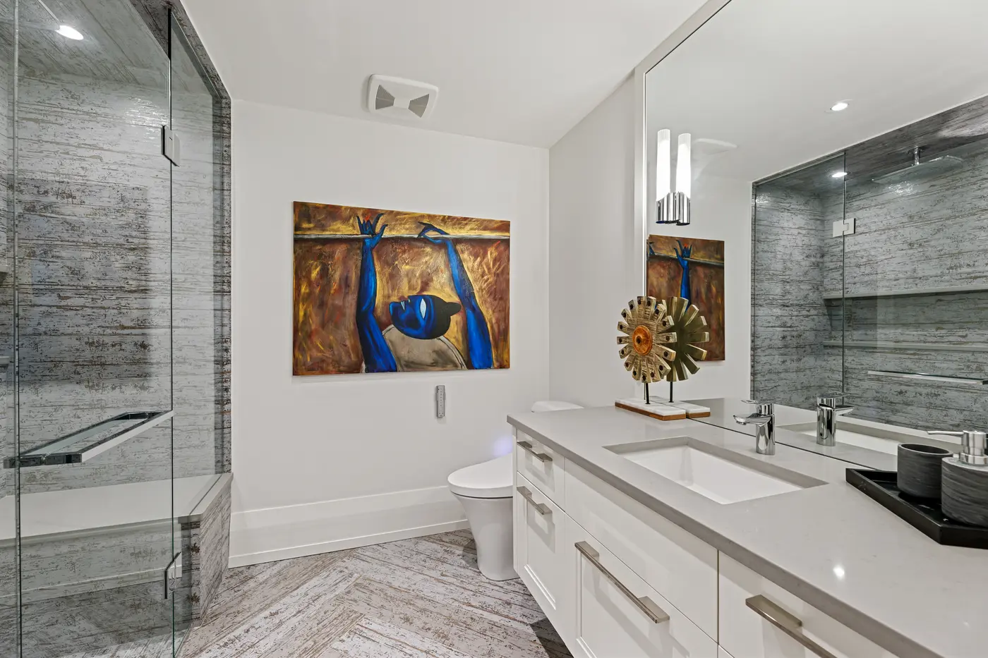 A lower-level bathroom layers rustic silver-toned plank tile in the glass shower against a quartz-topped floating vanity and a striking blue figurative canvas.