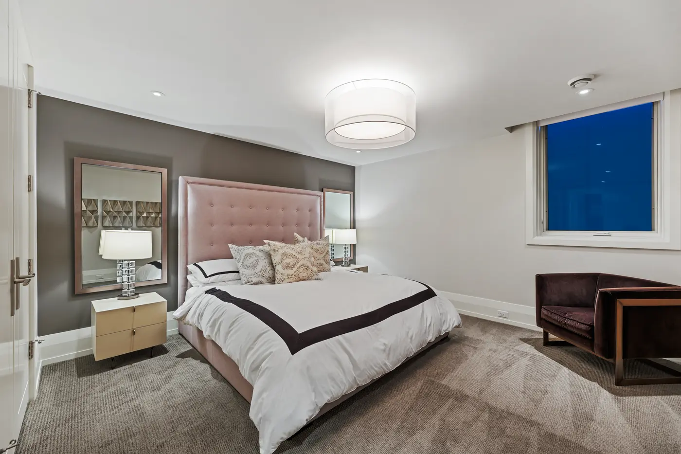 A serene guest bedroom with a blush tufted headboard wall, mirrored bedside vignettes and a velvet accent chair beneath a drum pendant.