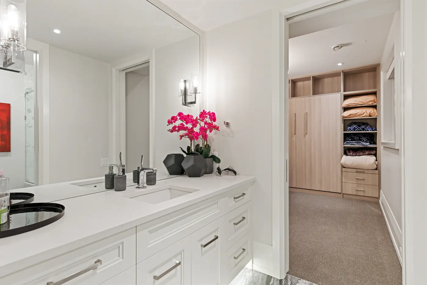A crisp white ensuite vanity with quartz counters and orchid styling opens through a pocket door to a fitted dressing room with warm oak built-ins.