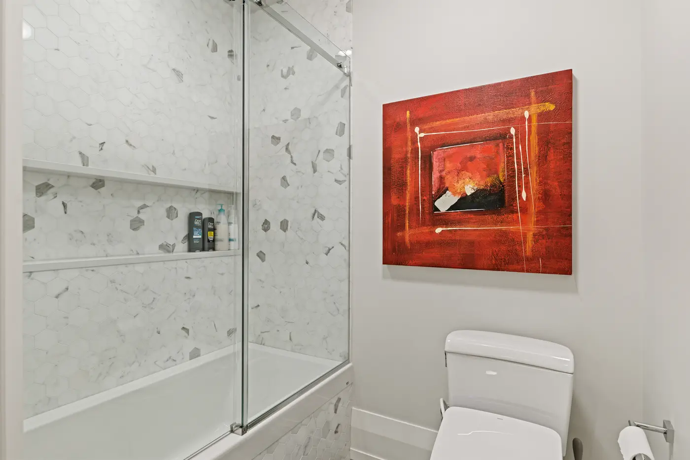 Marble hexagon mosaic wraps the tub-shower niche of a guest bath, warmed by a fiery abstract canvas above the Toto toilet.