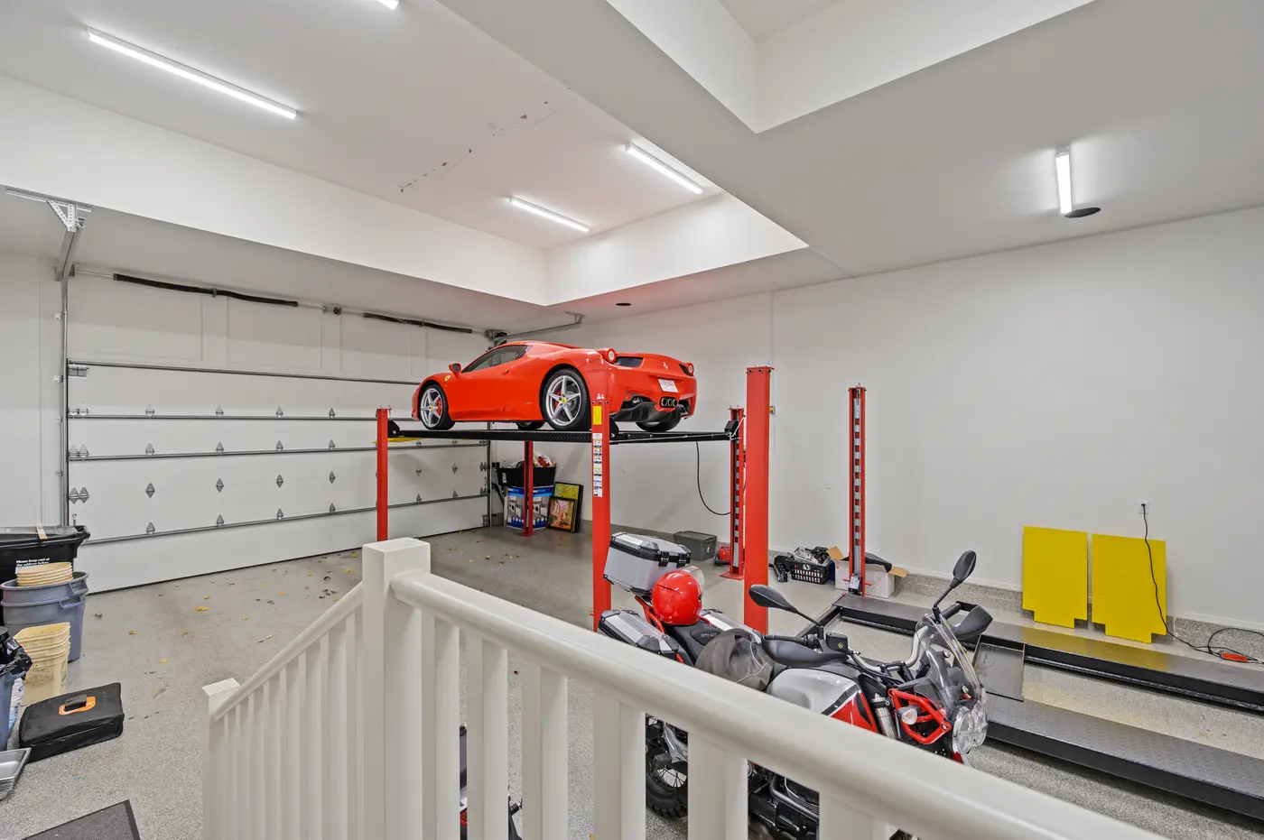 Inside the collector's garage, a red Ferrari 458 rests atop a four-post lift above a Moto Guzzi motorcycle and epoxy-flake flooring.