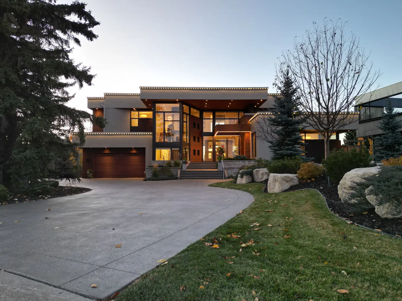 At dusk the estate glows from within, its glass-wrapped entry tower, warm walnut garage door and soffit lighting rising above the sweeping exposed-aggregate drive.