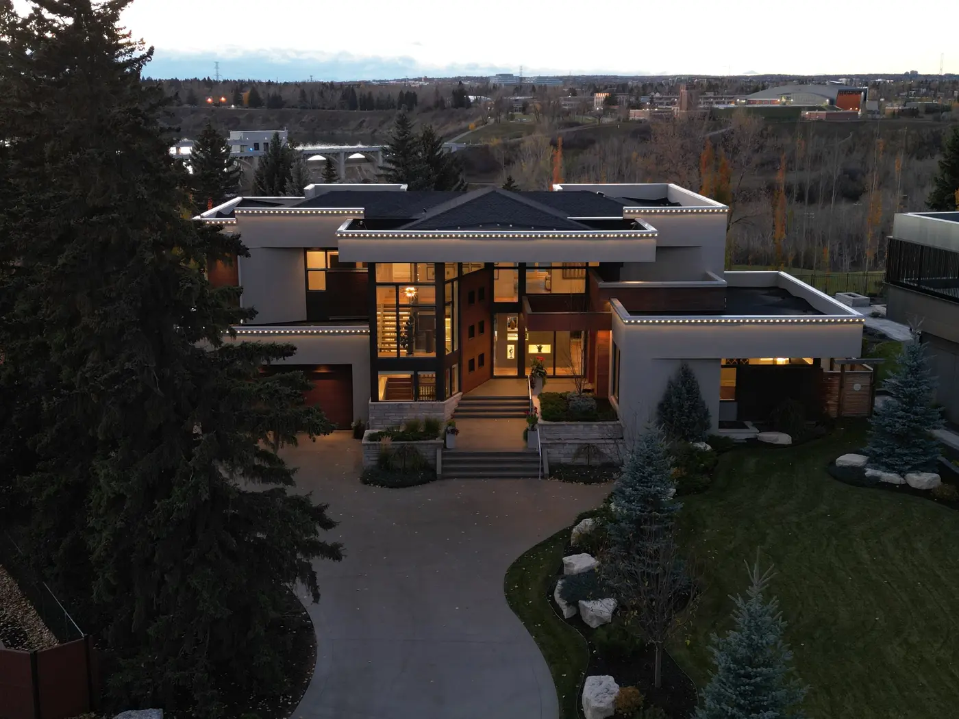 A twilight aerial over the front elevation reveals the home's layered flat rooflines and lantern-lit entry, with the Glenmore Reservoir, historic bridge and city skyline stretching behind.