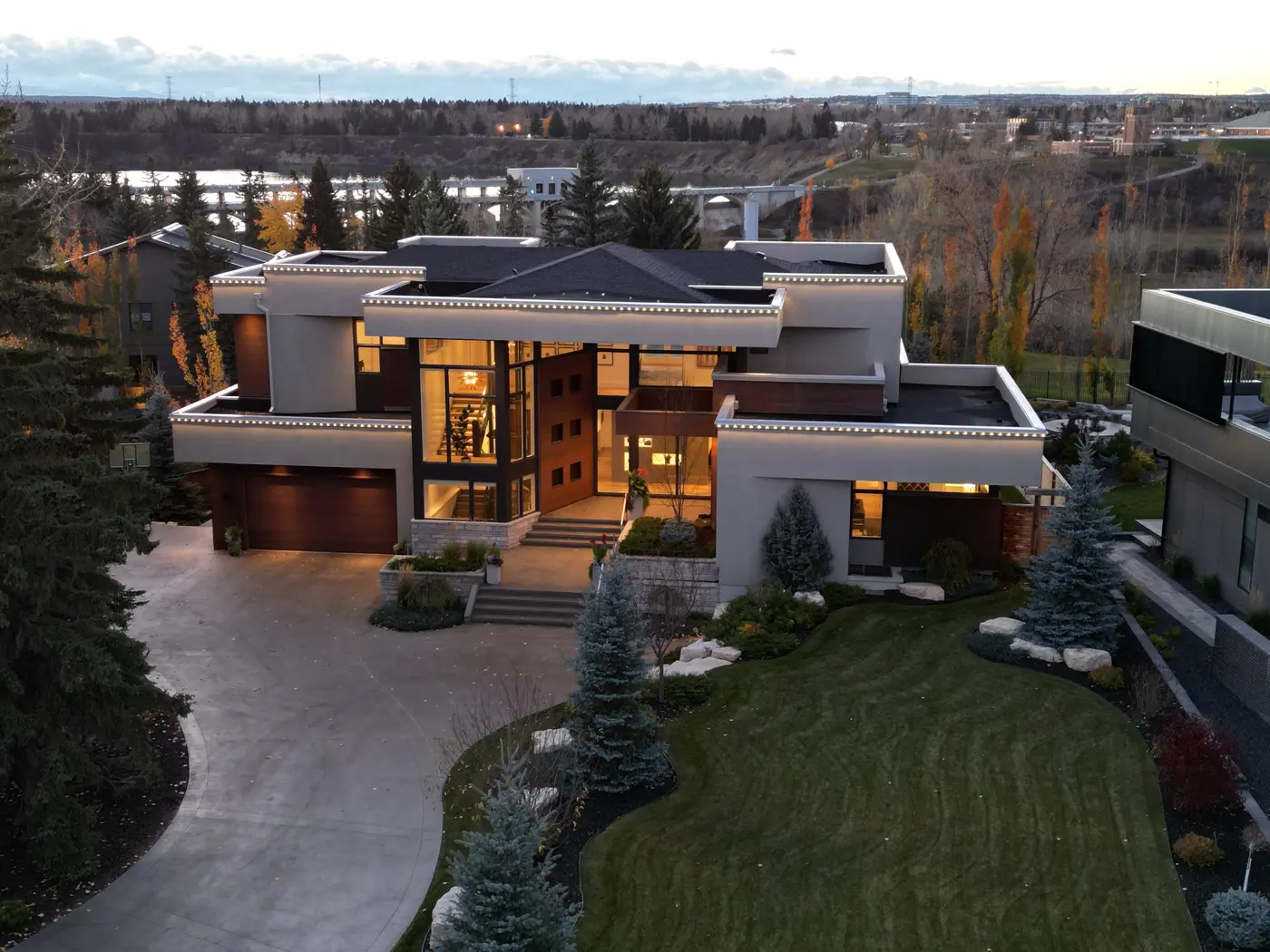 Hovering above the manicured front lawn at dusk, the drone captures the illuminated three-storey glass entry with the Glenmore weir bridge and reservoir shimmering beyond.