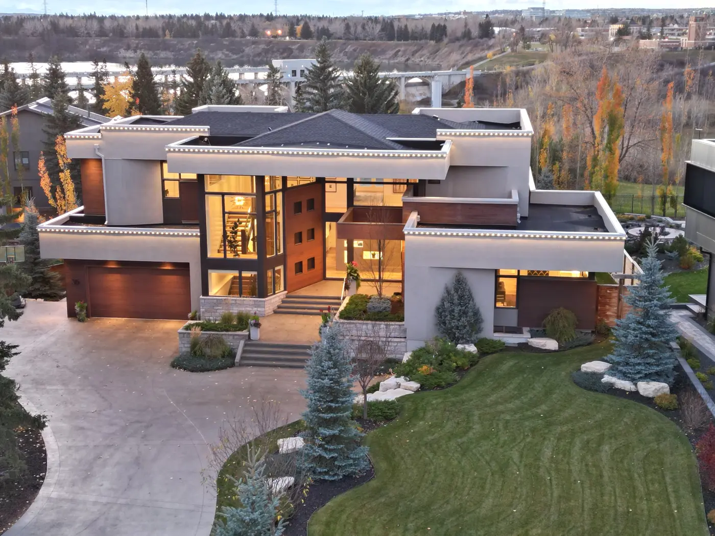 A closer twilight aerial of the facade showcases the floating rooflines traced in light, sculpted evergreens and stone-terraced entry stairs, with the reservoir bridge in the distance.