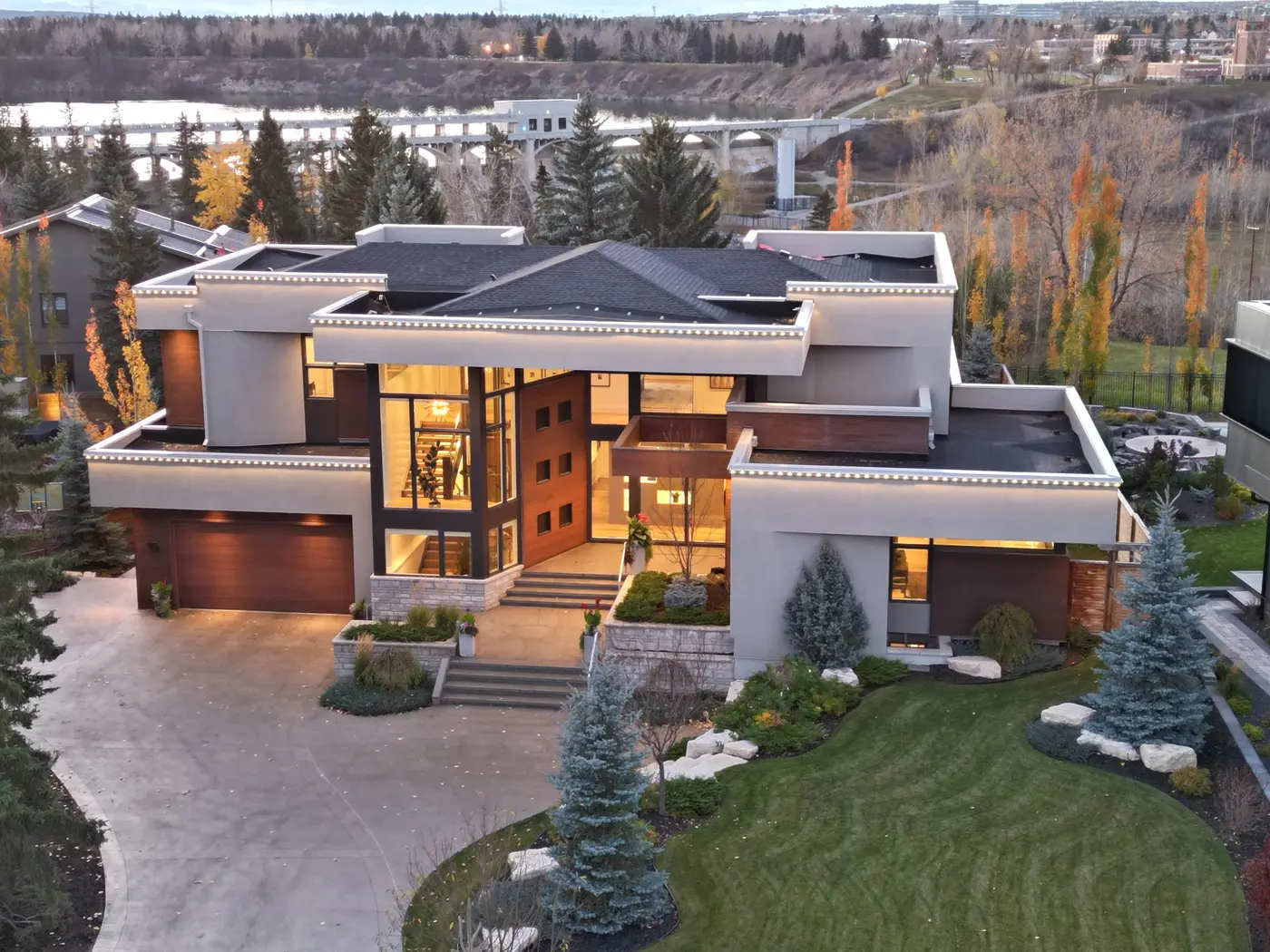 The estate's full front composition from the air at dusk, its glowing staircase tower and layered terraces set against the Glenmore Reservoir, weir bridge and autumn river valley.