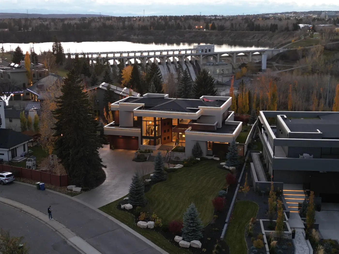 An elevated dusk view down the street frontage frames the home between mature spruce and the sweeping curve of the drive, with the Glenmore weir and reservoir beyond the treed escarpment.