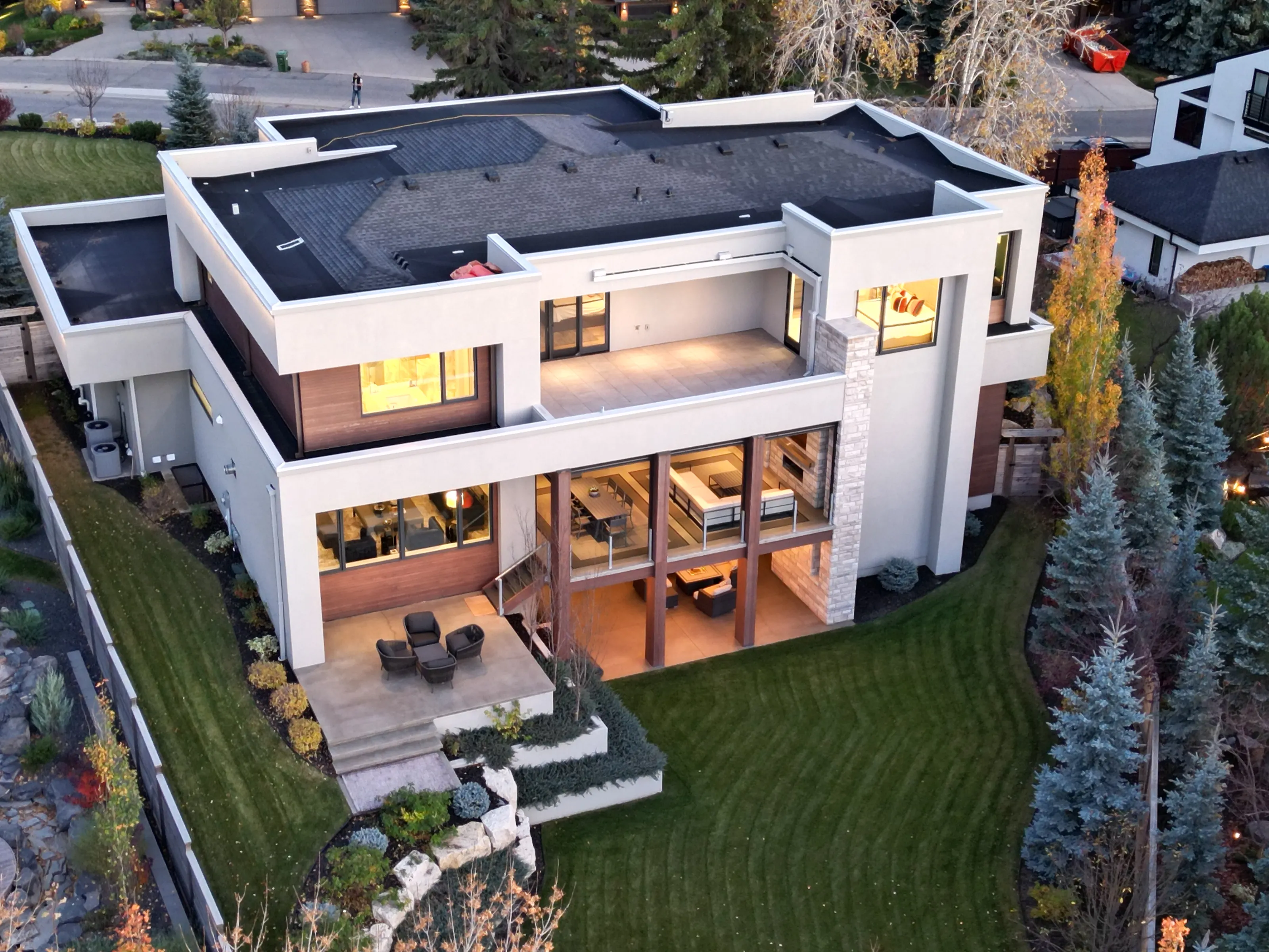 From above the rear elevation at dusk, the two-storey covered outdoor living room glows between stone columns, overlooking the terraced walkout patio and striped lawn.
