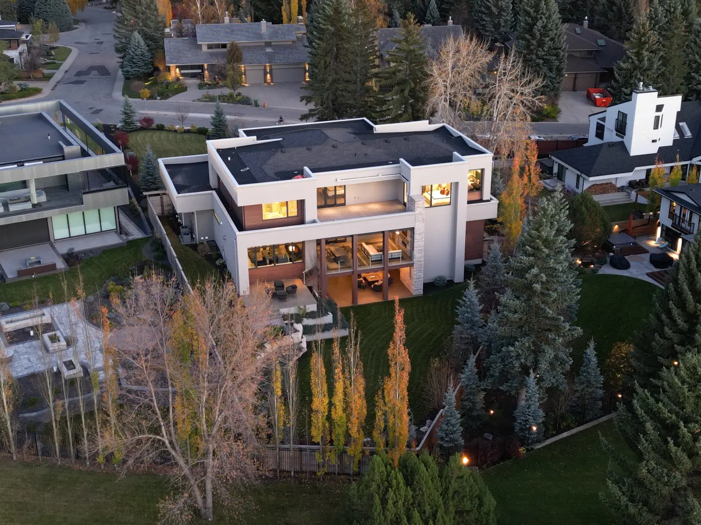 A rear aerial at twilight shows the walkout backyard descending through rock gardens and golden poplars toward the fenceline bordering the Glenmore parkland.