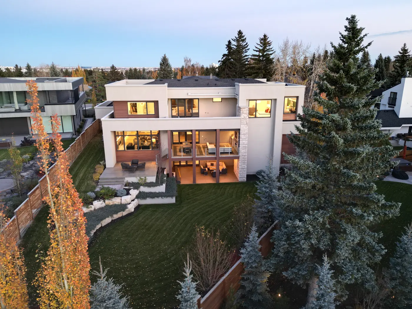The back of the home lit for evening, its double-height covered terrace and upper balcony overlooking the private lawn, with the city skyline on the horizon.
